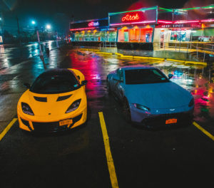 Yellow Lotus Evora and grey Aston Martin Vantage in front of neon lights in the rain